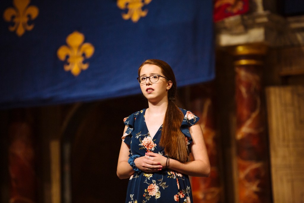 Emily Parise standing on the Globe Theatre stage in London, mid-performance. She wears a blue floral dress.